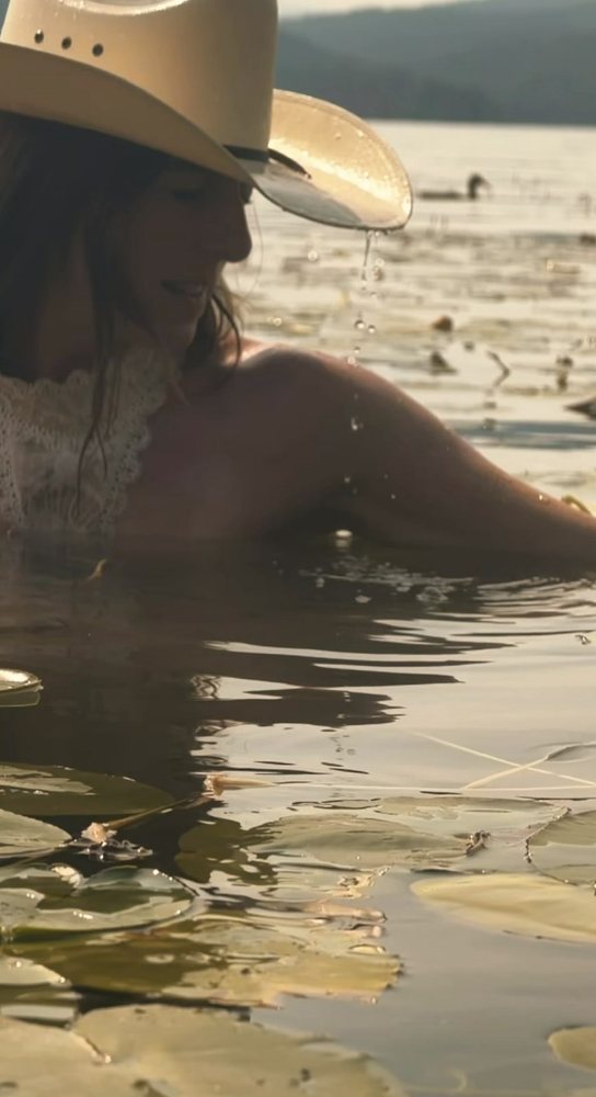 A woman in a cowboy hat, backlit, water dripping from the brim, among lily pads. Photograph by Hk.