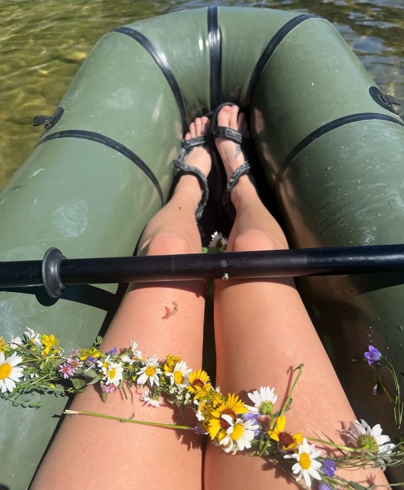 Bare legs in a green packraft, a wildflower garland and a paddle across the knees. Photograph by Hk.