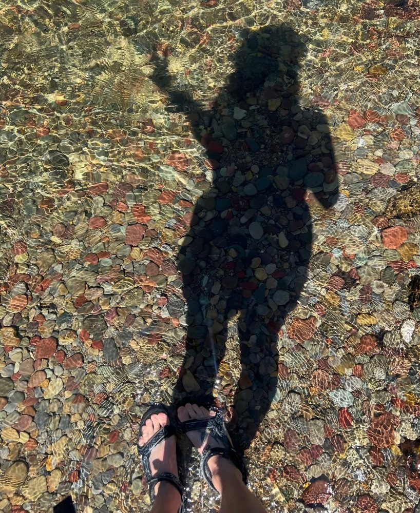 A person&rsquo;s shadow cast over colorful pebbles under clear, sunlit river water. Photograph by Hk.