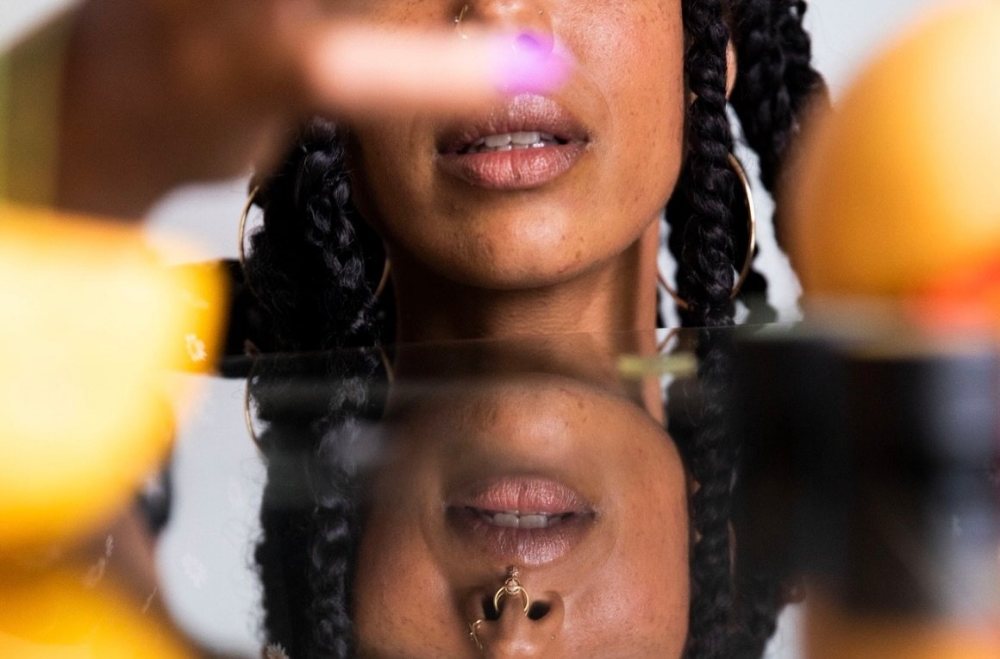 A close crop of lips and a nose ring mirrored on glass, citrus blurred in front. Photograph by Hk.