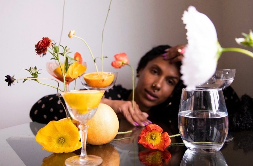A table of poppies, citrus halves in glasses and water, a woman resting behind it. Photograph by Hk.